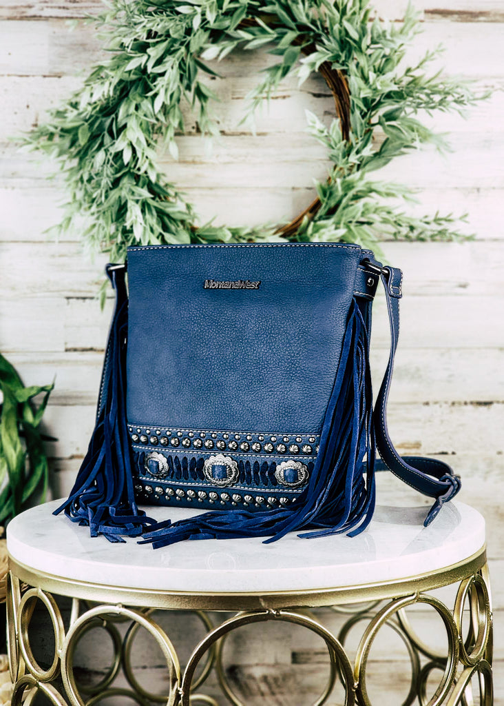 Concealed carry navy leather with accent silver conchos and studs. Blue accented stitching with silver concho buckles. Navy fringe hangs from the side of the purse with adjustable shoulder strap. Purse is displayed angled on a marble table top.
