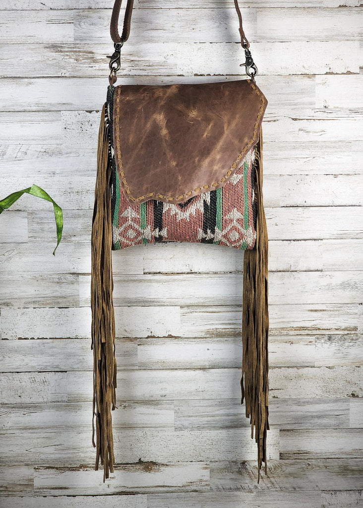 Concealed carry crossbody with leather flap snap with southwestern woven design. Fringe falls from the side of the purse. Purse hangs against a wooden wall. 