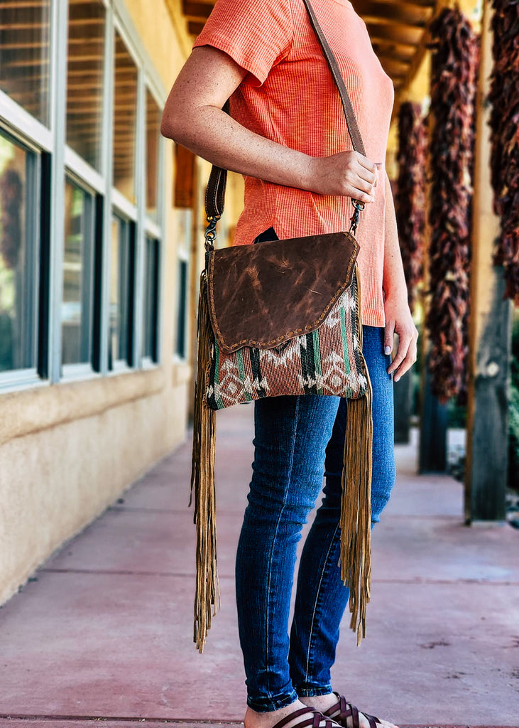 Concealed carry crossbody with leather flap snap with southwestern woven design. Fringe falls from the side of the purse. Model wears purse over her shoulder. 
