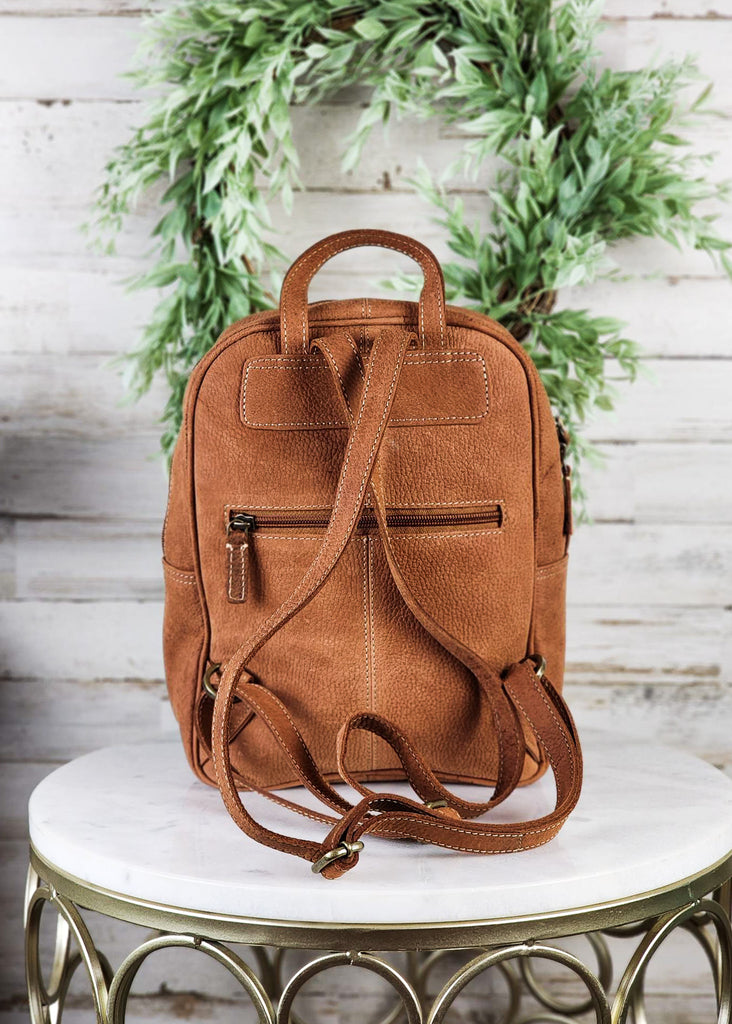 Backside of backpack is displayed. Back zipper small pocket is seen along with adjustable shoulder strap and top handle. Purse is displayed on marble table top.