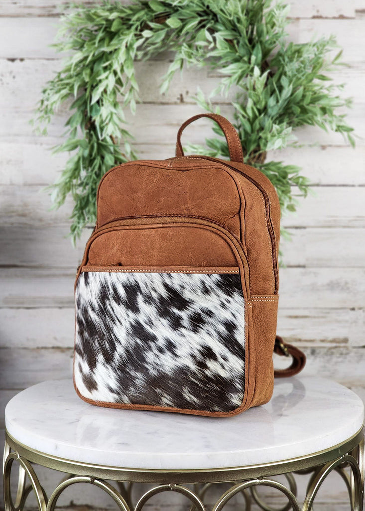 Cowhide pocket panel with medium oil leather. Cowhide and leather backpack has two exterior zipper pockets and side open pockets and back pocket on the cowhide. Purse is displayed on marble table top. 