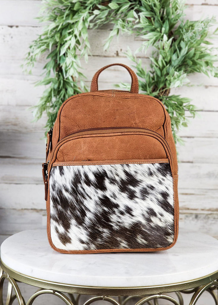 Cowhide pocket panel with medium oil leather. Cowhide and leather backpack has two exterior zipper pockets and side open pockets and back pocket on the cowhide. Backpack is displayed on marble table top. 