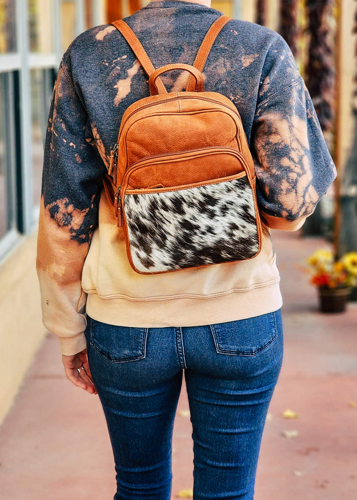 Cowhide pocket panel with medium oil leather. Cowhide and leather backpack has two exterior zipper pockets and side open pockets and back pocket on the cowhide. Model wears this over her back showing the shoulder straps and zippered pockets.