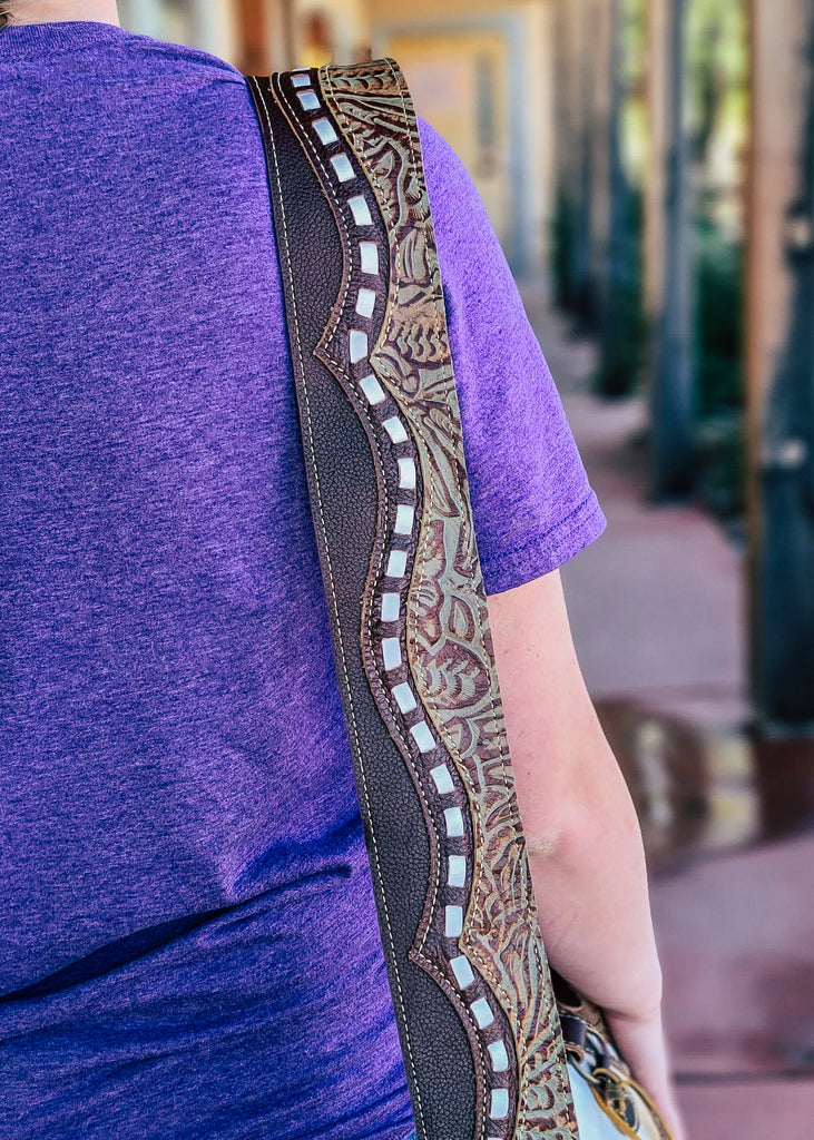 Brown and white cowhide crossbody handbag with floral tooled embossed leather accented detail on the sides with brown fringe on the sides, with steer skull on the front, taken outside on model 