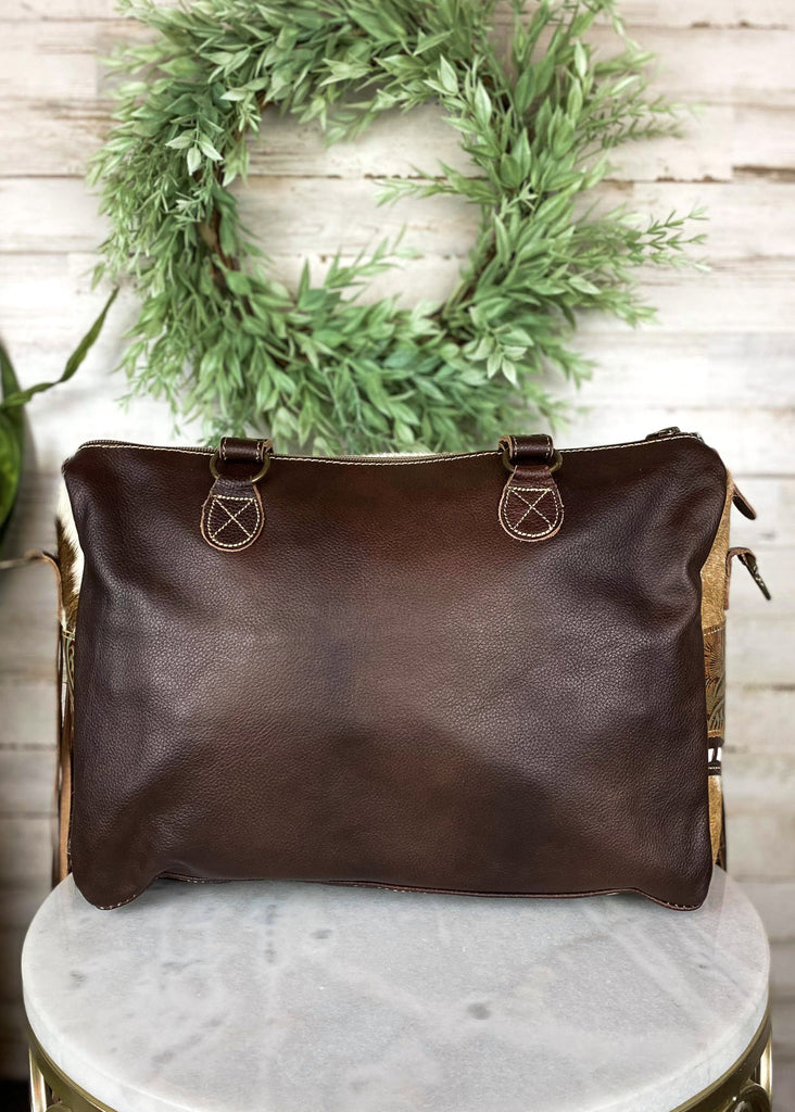 Brown and white cowhide crossbody handbag with floral tooled embossed leather accented detail on the sides with brown fringe on the sides, with steer skull on the front, taken inside on white table with green floral décor 