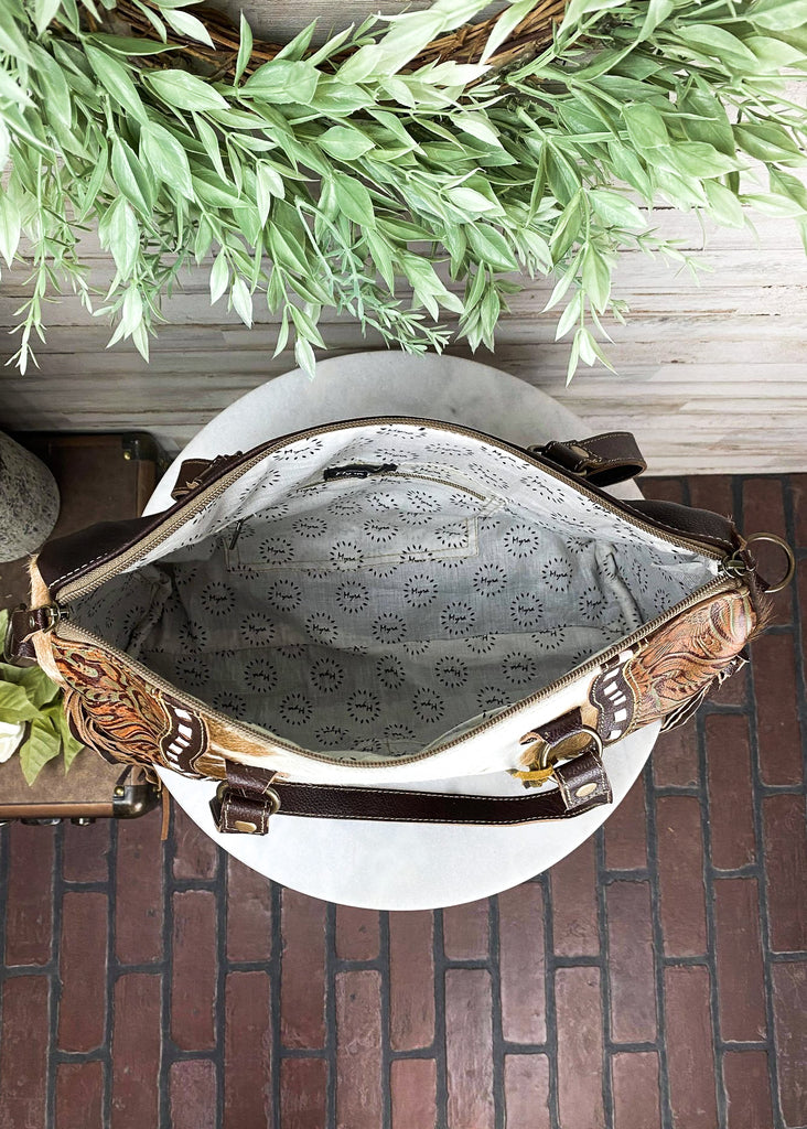 Brown and white cowhide crossbody handbag with floral tooled embossed leather accented detail on the sides with brown fringe on the sides, with steer skull on the front, taken inside on white table with green floral décor 