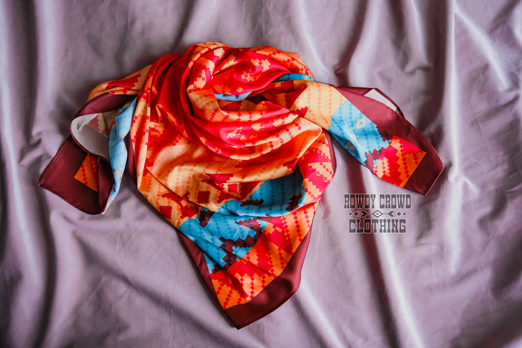 Western wild rag with bright reds and orange tribal design and two strips of turquoise and maroon. Wild rag coiled to imitate who it would look on a neck. 