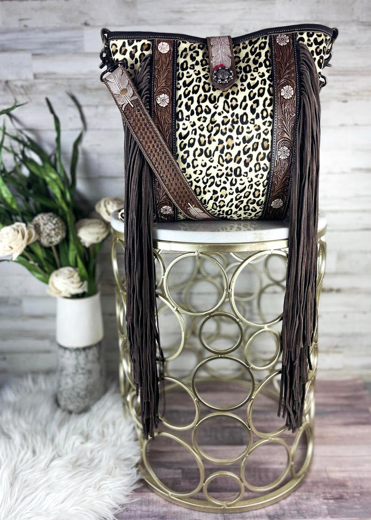 Leaopard print purse with floral tooled leather accent strips run up the purse with long fringe. Matching shoulder straps with feather tooling. Purse sits on marble table top. 