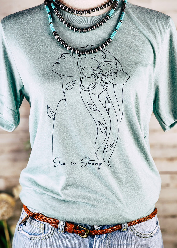 Dusty blue short sleeve with black outline of a women and black lettering reading "She is strong". Tee hangs on mannequin with chocker necklace 