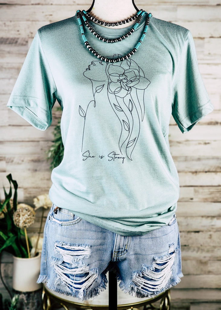 Dusty blue short sleeve with black outline of a women and black lettering reading "She is strong". Tee hangs on mannequin with chocker necklace and jean shorts.