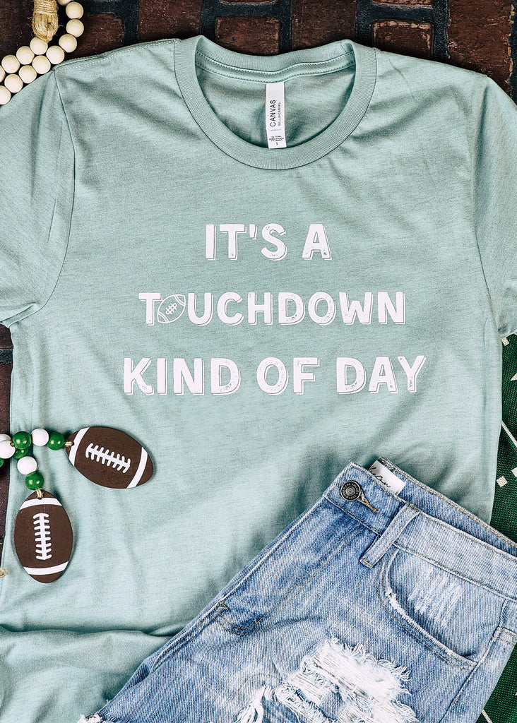 Dusty blue short sleeve tee with white lettering reading "It's a touchdown kind of day". The tee lays on a brick floor with jean shorts and football decor.