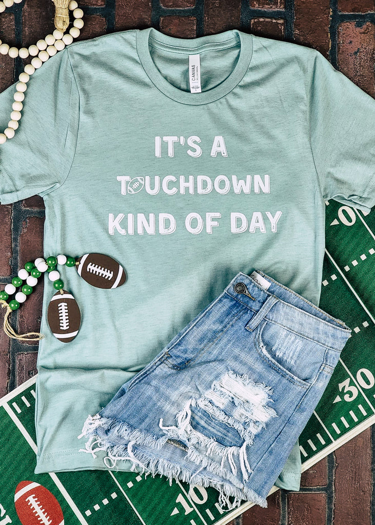 Dusty blue short sleeve tee with white lettering reading "It's a touchdown kind of day". The tee lays on a brick floor with jean shorts and football decor. 