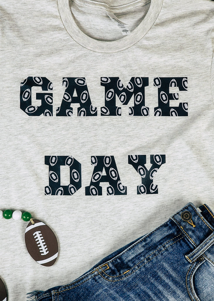 Heather natural short sleeve with black lettering reading "Game Day" with white outlined footballs on the lettering. Football decor and jeans decorate the tee. 