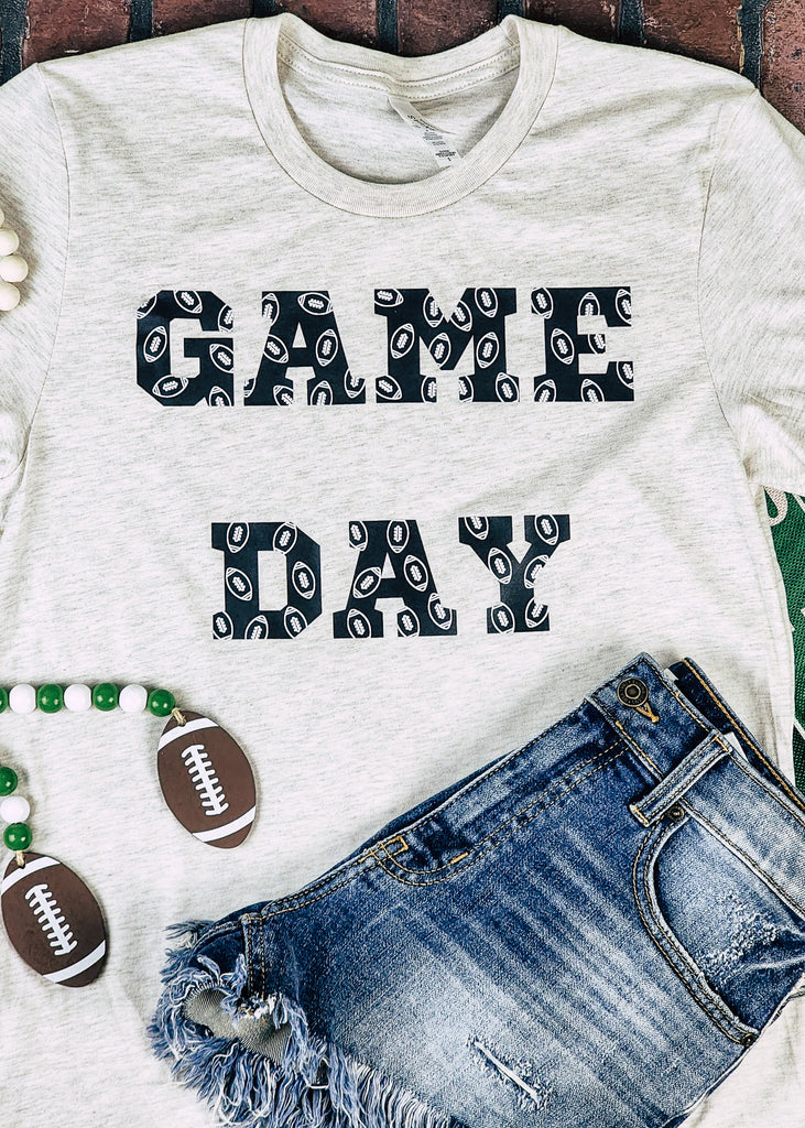Heather natural short sleeve with black lettering reading "Game Day" with white outlined footballs on the lettering. Tee lays on brick background with football decor and jean shorts.