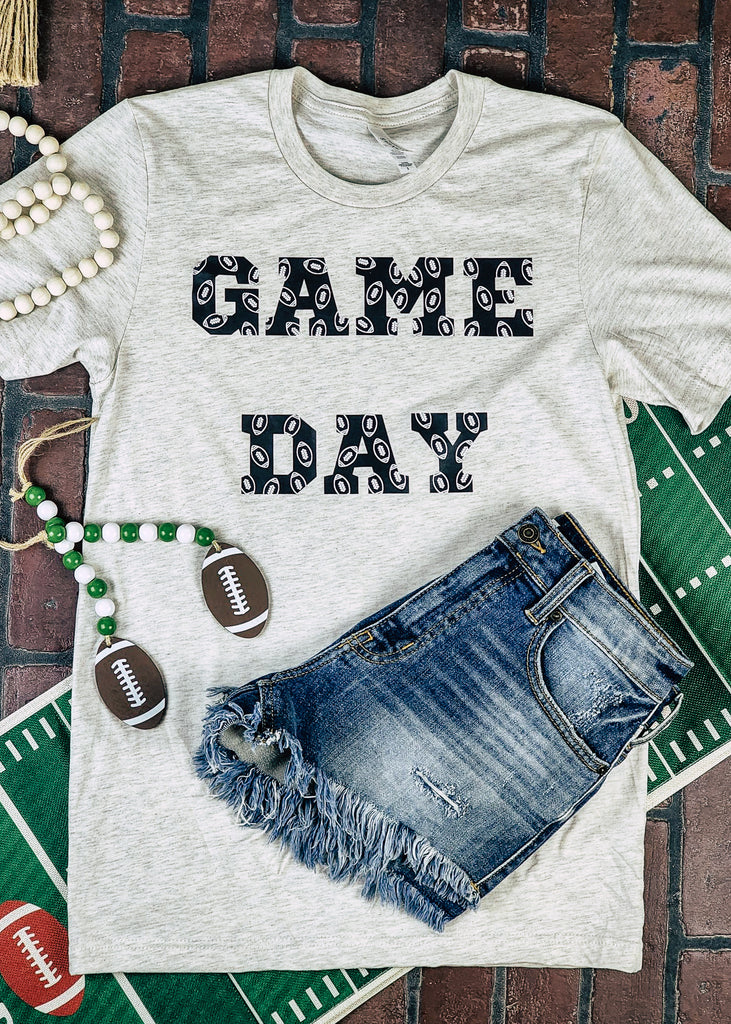 Heather natural short sleeve with black lettering reading "Game Day" with white outlined footballs on the lettering. Tee lays on brick background with football decor and jean shorts.