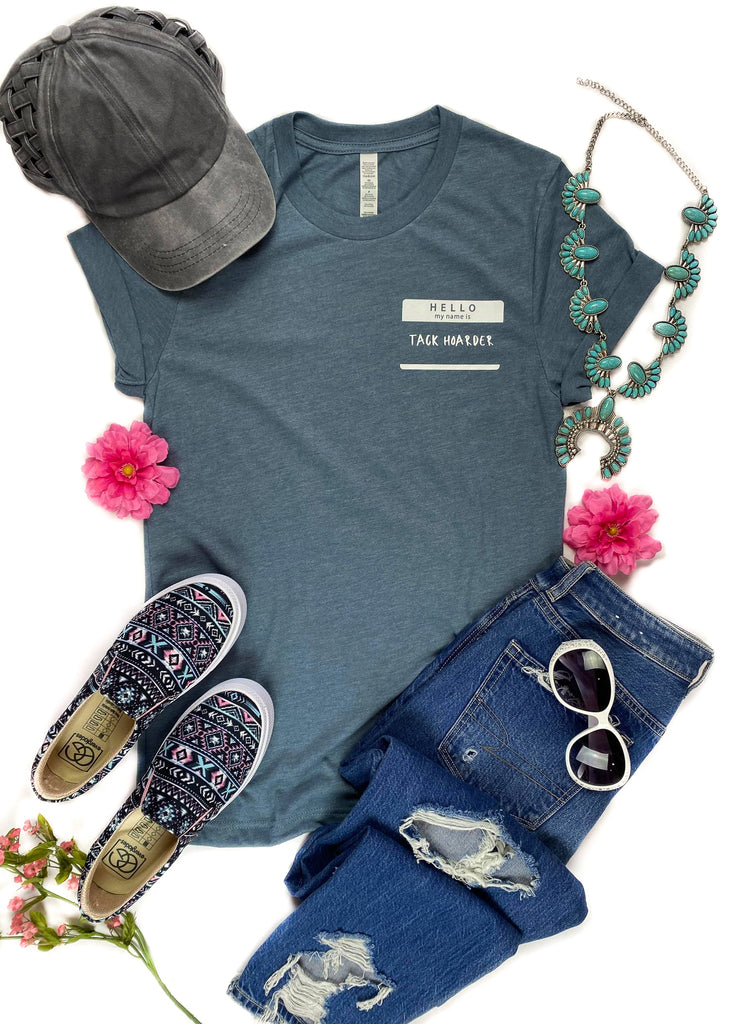 Heather slate colored short sleeve tee with sleeves rolled up and name tag graphic in the right (left on person) that says "Hello my name is Tack Hoarder" in white ink, laid flat on white surface with denim pants, baseball cap, Aztec slip on shoes and turquoise squash blossom, with pink floral décor 