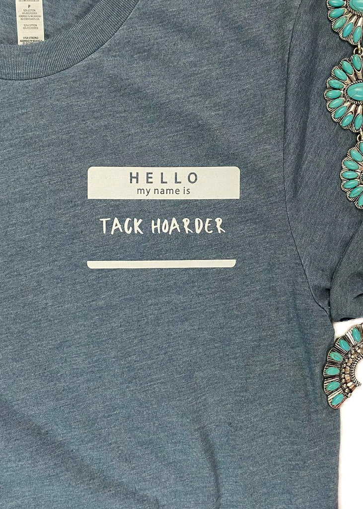 Heather slate colored short sleeve tee with sleeves rolled up and name tag graphic in the right (left on person) that says "Hello my name is Tack Hoarder" in white ink, laid flat on white surface with denim pants, baseball cap, Aztec slip on shoes and turquoise squash blossom, with pink floral décor 