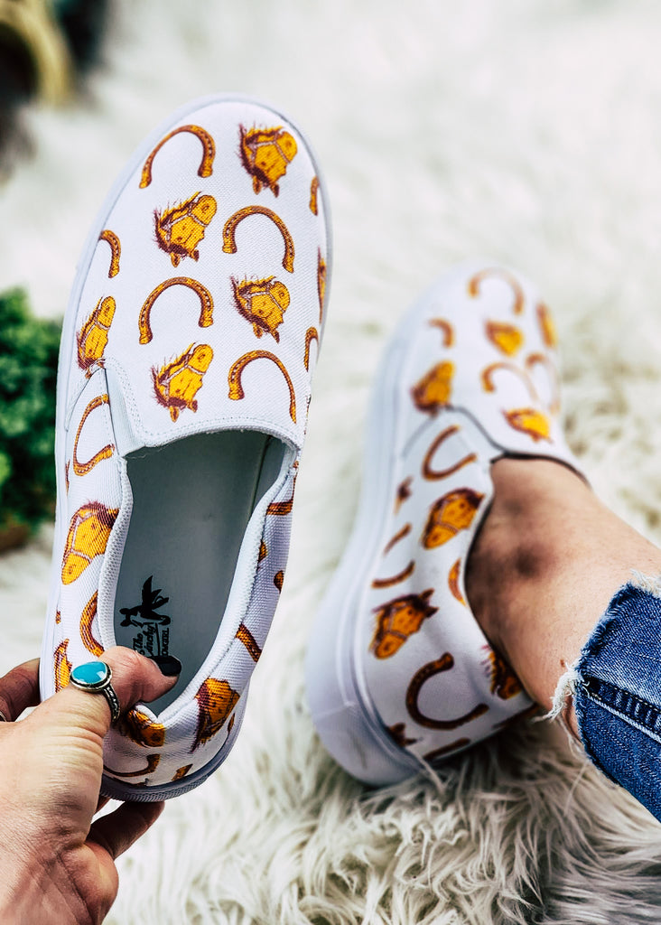 Horse heaven slip on shoes with white fabric, gold horse heads and horse shoe pattern. Model wears one shoe and holds the other against a white faux fur. 
