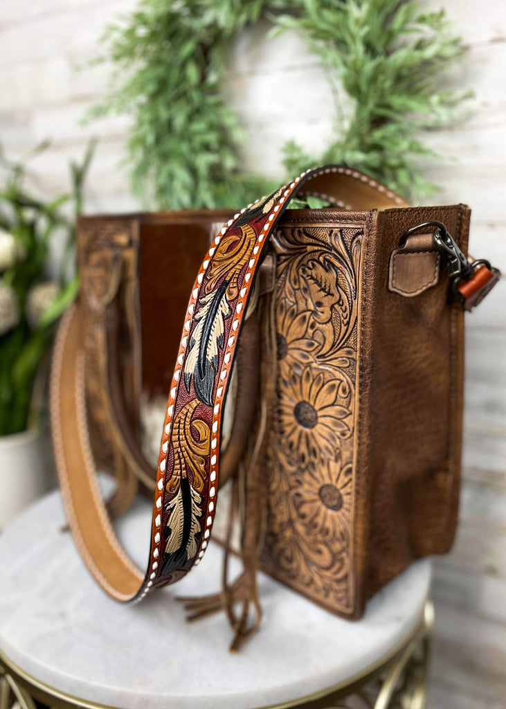 Multi colored purse strap with detailed feather with whipstitched detail, taken inside on brown handbag with floral décor 