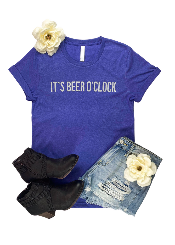 CLOSEOUT- Lapis short sleeve tee with grey lettering reading "It's beer o'clock". Tee is laid on white background with black booties, jean shorts, and white floral decor. 
