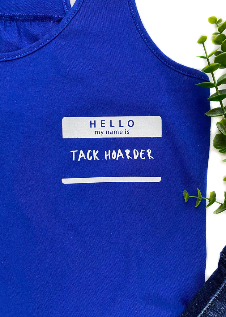 Royal Blue colored tank top with name tag graphic in the right (left on person) that says "Hello my name is Tack Hoarder" in white ink, laid flat on white surface with denim pants, black and gold sneakers, black baseball hat and floral décor 