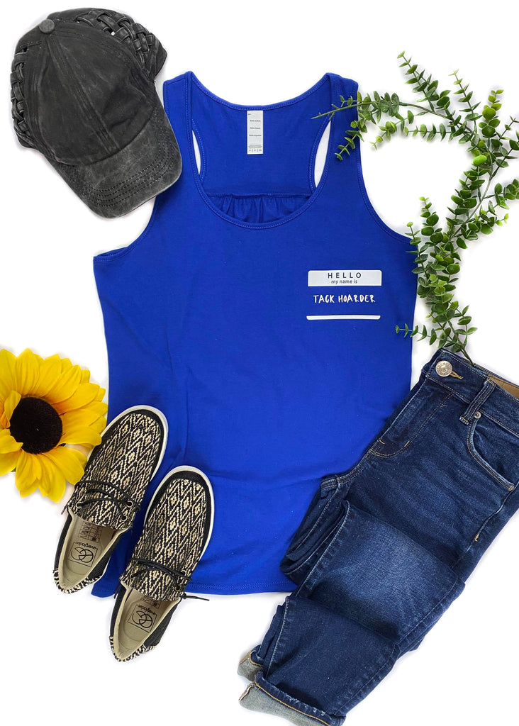 Royal Blue colored tank top with name tag graphic in the right (left on person) that says "Hello my name is Tack Hoarder" in white ink, laid flat on white surface with denim pants, black and gold sneakers, black baseball hat and floral décor 