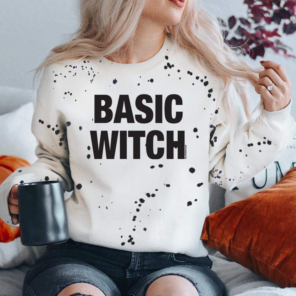 Western crewneck with black "blood splatter" with black font that reads "Basic Witch". Model is holding a black coffee mug surounded by burnt orange and white fall style pillows. 