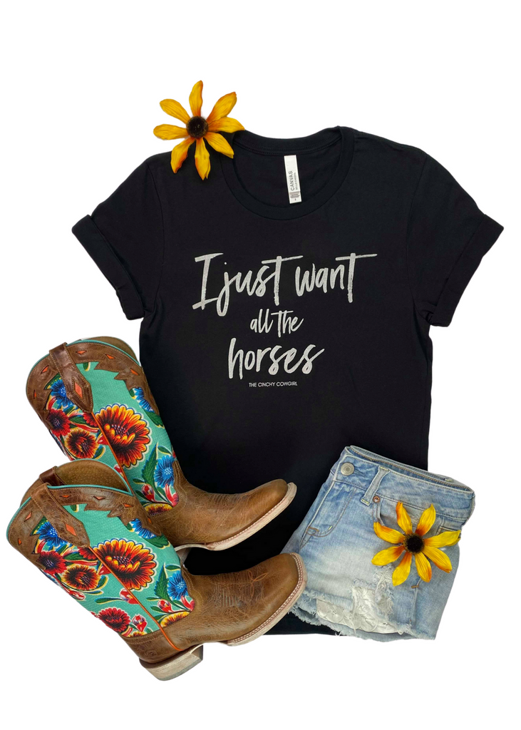 Black short sleeve tee with gray lettering reading "I just want all the horses". Shirt laying flat with sunflowers, floral boots, and jean shorts. 