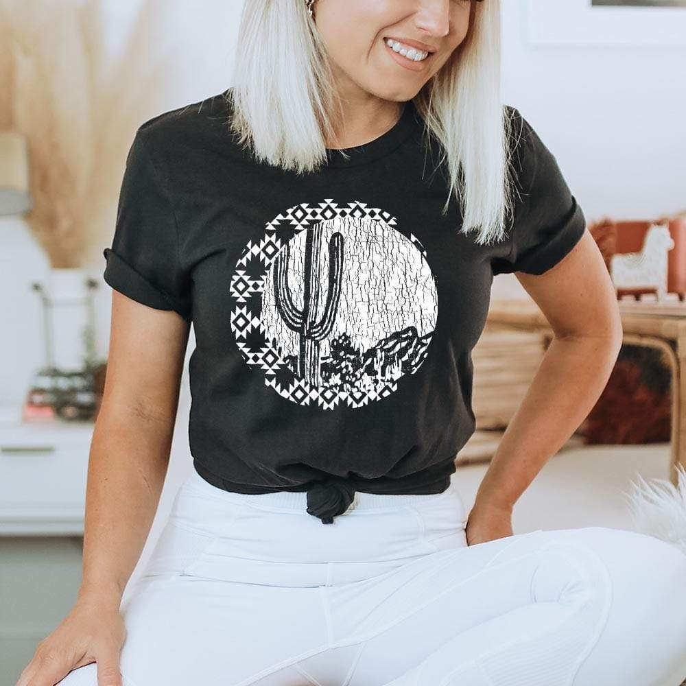 Black short sleeve tee with white printed southwestern crescent moon and white circle surrounded by crescent moon. Black negative space shows cracks and a night desert scene of a cactus close up and mountains. 