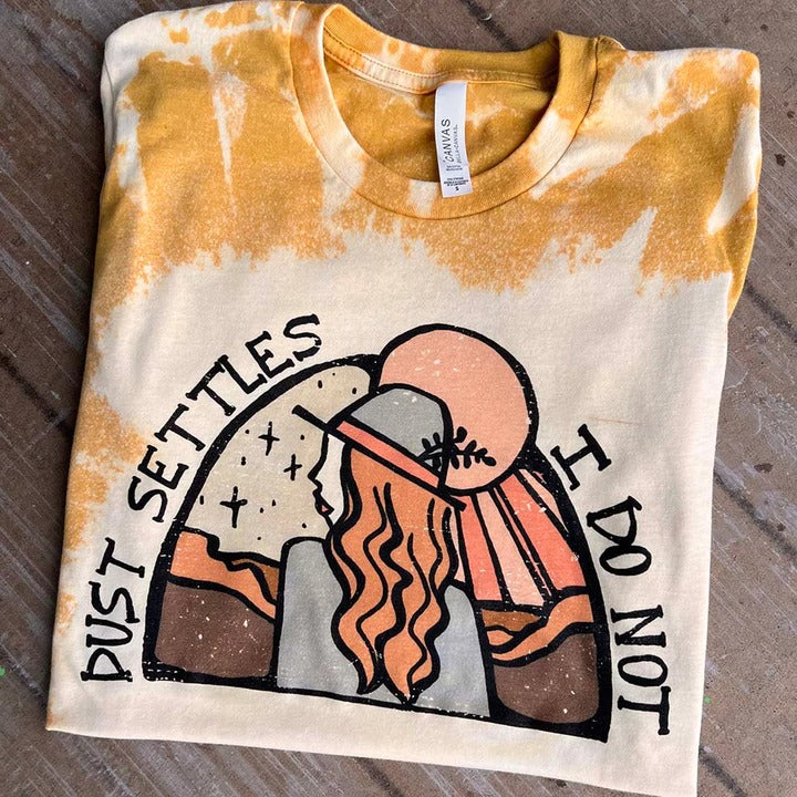 Bleached mustard short sleeve tee with black lettering reading "Dust settles I do not" with a desert sunset and cowgirl graphic. Tee is folded and lays on a wooden background. 