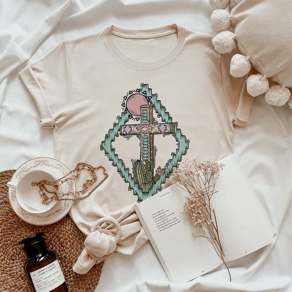 Cream short sleeve tee with aztec blue frame and an Easter cross scenery. Tee lays tied with the sleeves rolled on a white and cream theme background. 