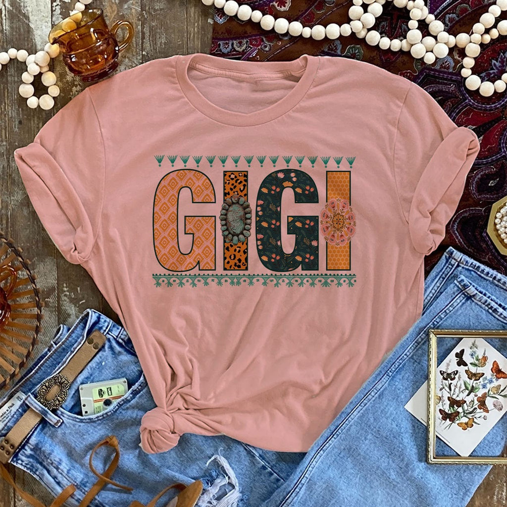 Desert rose short sleeve tee with Multi-pattern lettering reading "GIGI" each "I" on the conchos. Tee has the sleeves rolled with it tied at the bottom. Tee lies on the wood background. 