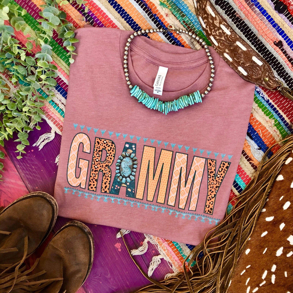 Mauve short sleeve tee with multi pattern lettering reading "GRAMMY". On the "A" there is a turquoise concho. Tee lays folded on purple wood with multi color rug with western decor. 