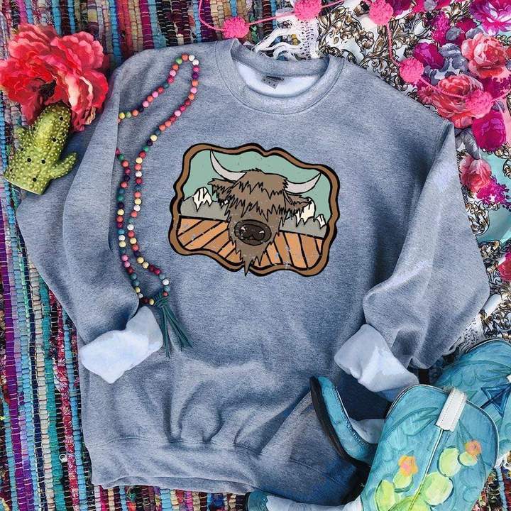 Gray crewneck with a highland cow with mountain background. Crewneck has the sleeves rolled with a colorful rug background. 