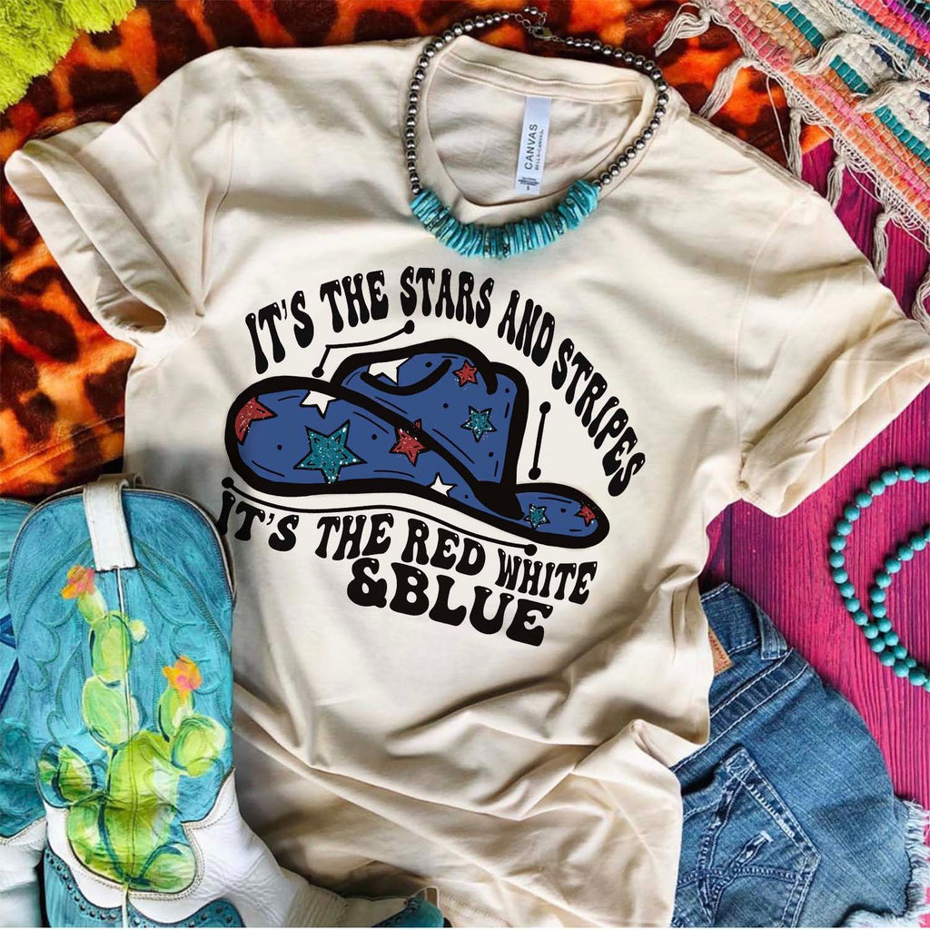 Cream short sleeve tee with black bubble lettering "It's the stars and stripes it's the red white and blue". A blue cowboy hot with red white and blue stars. The tee has its sleeves rolled while laying on a pink wooden background. 