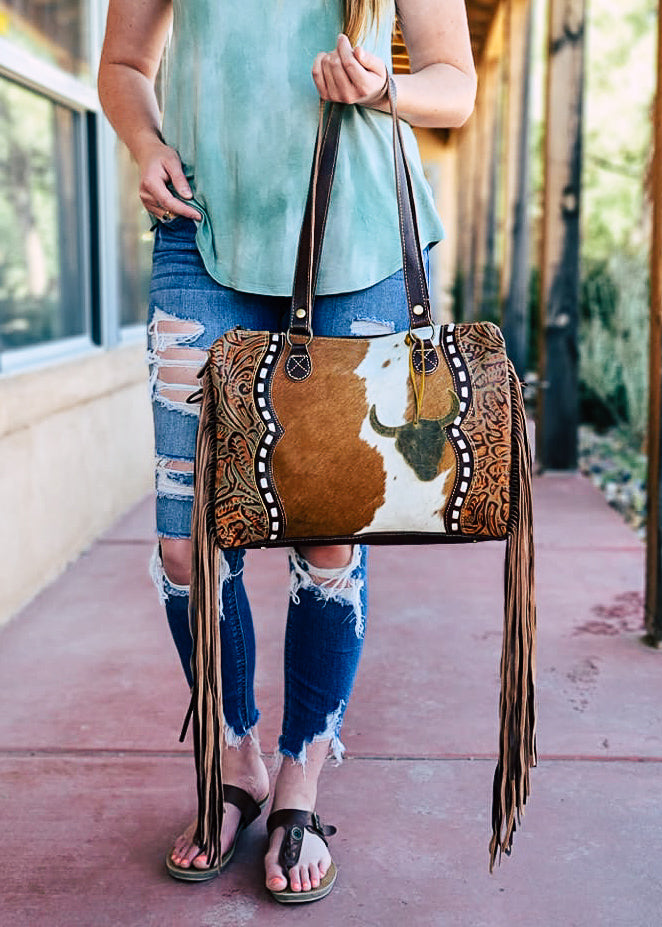 Western crossbody bag with cowhide inlay with tooled leather side panels. White whip stitch lines the leather panels. Steer skull cutout is attached to handle straps. Fringe hangs from the side of the bag. Model wears this over her shoulder with matching tooled and whip stitch shoulder strap. Stye A