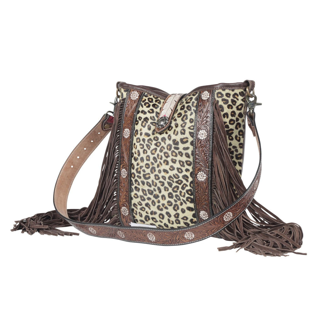 Leaopard print purse with floral tooled leather accent strips run up the purse with long fringe. Matching shoulder straps with floral tooling. Purse sits on marble table top. 