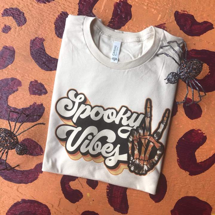 Cream short sleeve tee with bubble cursive lettering reading "Spooky Vibes" with Halloween colored shadows and tie-die skeleton peace sign. 