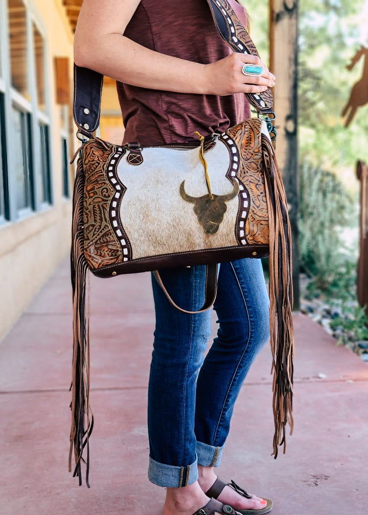  Western crossbody bag with cowhide inlay with tooled leather side panels. White whip stitch lines the leather panels. Steer skull cutout is attached to handle straps. Fringe hangs from the side of the bag. Model wears this over her shoulder with matching tooled and whip stitch shoulder strap. Style A