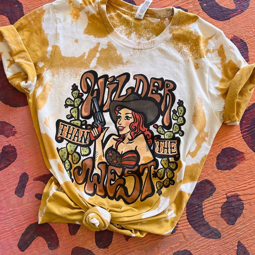 Bleached mustard short sleeve tee with bubble letters reading "Wilder than the West" with a cowgirl holding a pistol and cactus on the sides. Tee is tied at the bottom and sleeves are rolled up. 