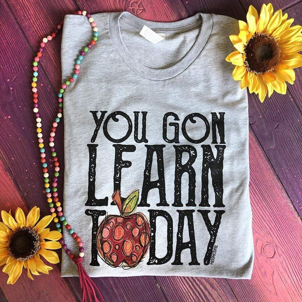 Grey short sleeve tee with black lettering reading "You Gon Learn Today" with an apple design acting as the "O" in the word today. Tee is folded on a pink background with sunflower decor. 